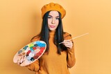 Young brunette woman holding paintbrush and palette wearing beret relaxed with serious expression on face. simple and natural looking at the camera.