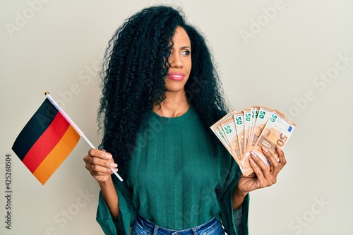 Middle age african american woman holding germany flag and euros banknotes smiling looking to the side and staring away thinking.