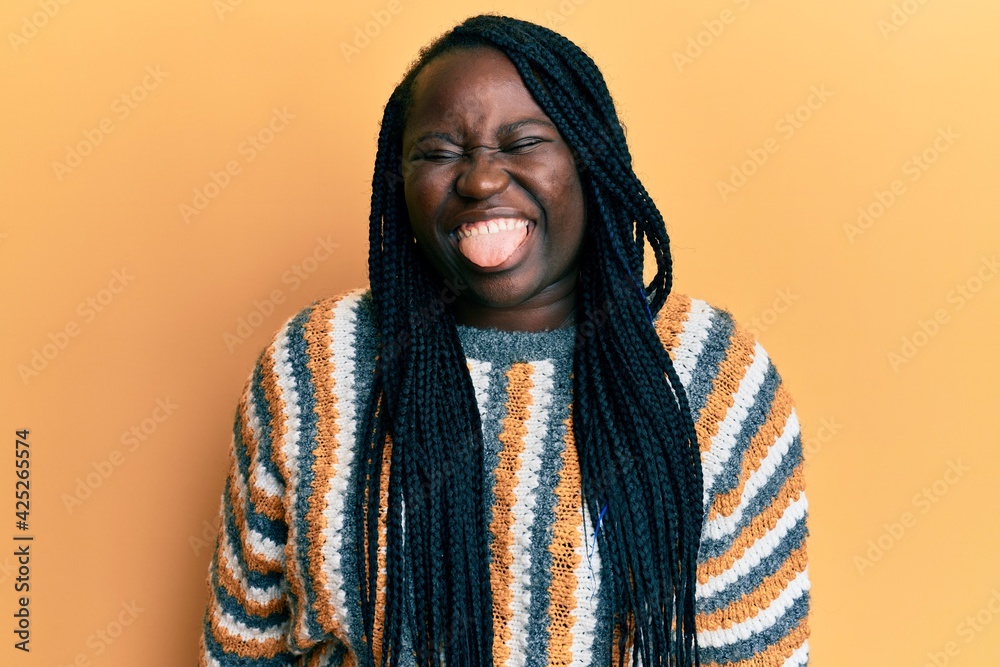 Young black woman with braids wearing casual winter sweater sticking tongue out happy with funny expression. emotion concept. Young black woman with braids wearing casual winter sweater sticking tongue out happy with funny expression. emotion concept.