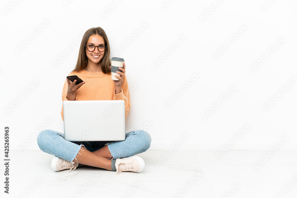 Young woman with a laptop sitting on the floor holding coffee to take away and a mobile Young woman with a laptop sitting on the floor holding coffee to take away and a mobile