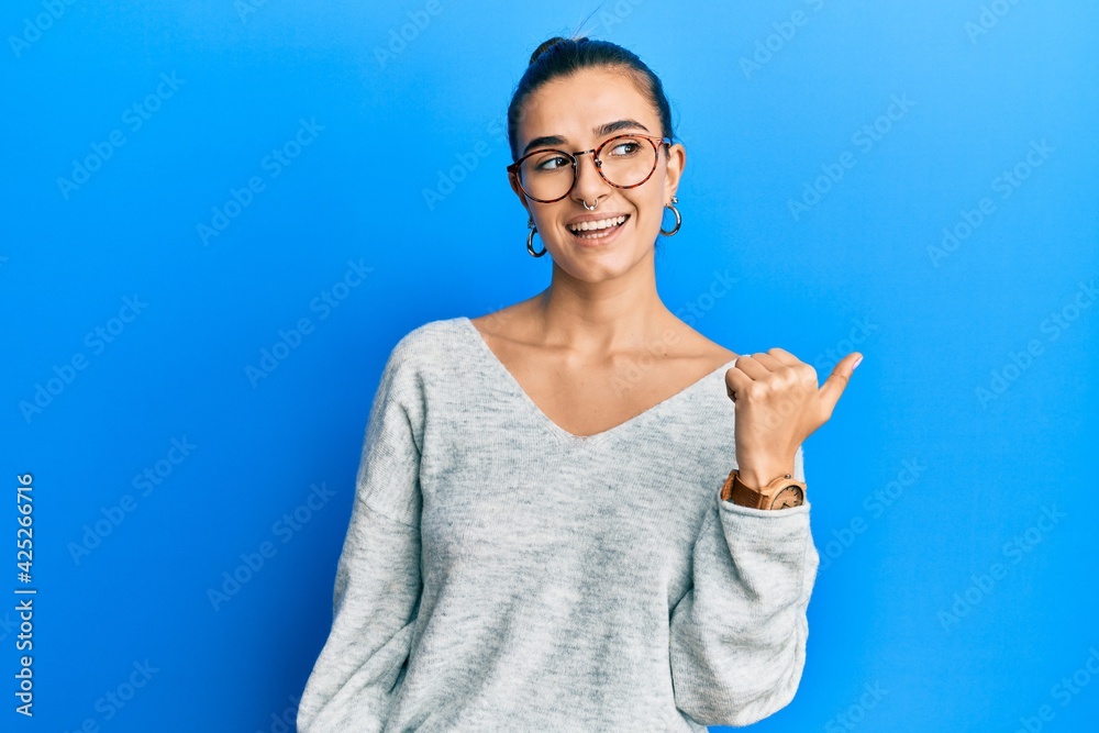 Young hispanic woman wearing casual clothes smiling with happy face looking and pointing to the side with thumb up. Young hispanic woman wearing casual clothes smiling with happy face looking and pointing to the side with thumb up.