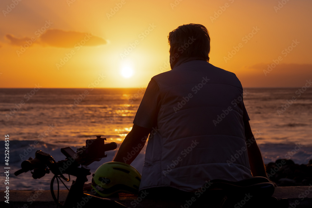Rear view of mature man in front of a magnificent orange sunset at sea. Bicycle by his side