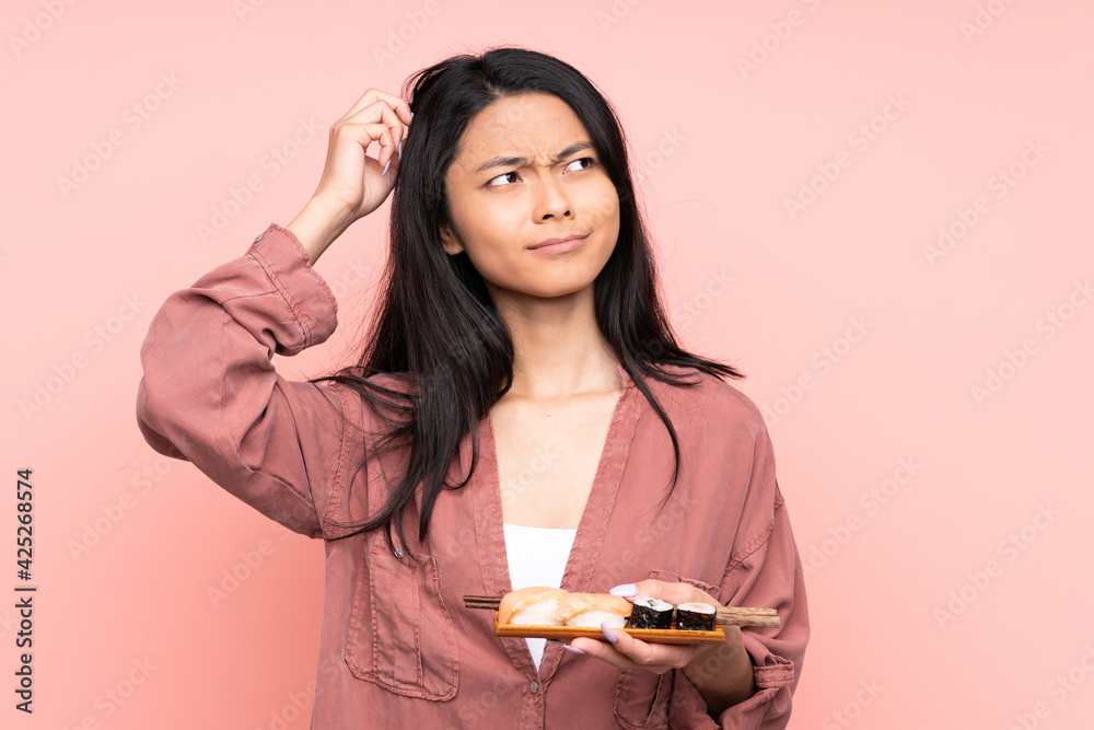 Teenager Asian girl eating sushi isolated on pink background having doubts and with confuse face expression Teenager Asian girl eating sushi isolated on pink background having doubts and with confuse face expression