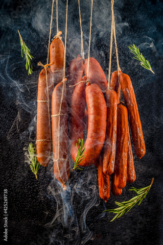 Smoking vertically hanging sausages. 
Clouds of smoke rise up and envelop the sausages hanging in a row. Dark background with flying green rosemary leaves