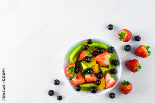 Sliced fresh ripe fruit  strawberries  kiwis  citrus fruits  blueberries  on a white plate. Whole large ripe strawberries and blueberries around the plate. White background.