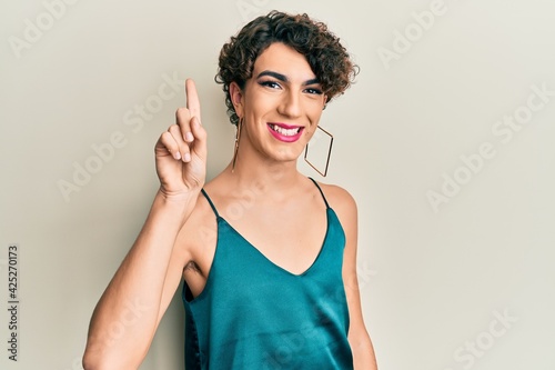 Young man wearing woman make up wearing party clothes smiling with an idea or question pointing finger up with happy face, number one