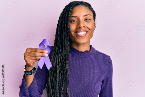 African american woman holding purple ribbon awareness looking positive and happy standing and smiling with a confident smile showing teeth photo