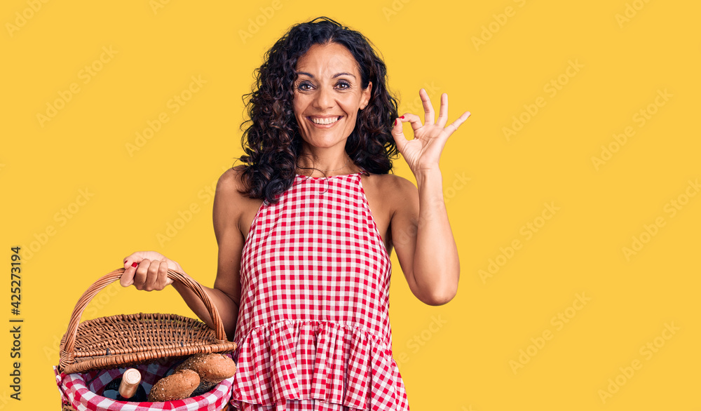 Middle age beautiful woman holding picnic basket with food doing ok sign with fingers, smiling friendly gesturing excellent symbol Middle age beautiful woman holding picnic basket with food doing ok sign with fingers, smiling friendly gesturing excellent symbol