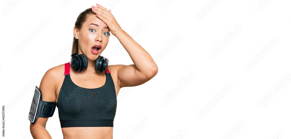 Beautiful blonde woman wearing gym clothes and using headphones surprised with hand on head for mistake, remember error. forgot, bad memory concept. Beautiful blonde woman wearing gym clothes and using headphones surprised with hand on head for mistake, remember error. forgot, bad memory concept.