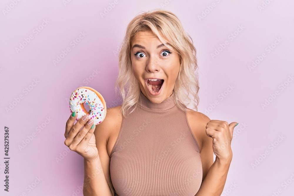 Young blonde girl holding donut pointing thumb up to the side smiling happy with open mouth Young blonde girl holding donut pointing thumb up to the side smiling happy with open mouth
