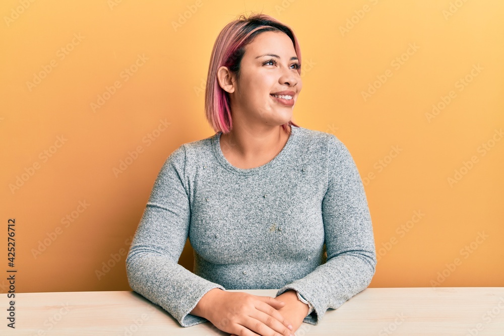 Hispanic woman with pink hair wearing casual clothes sitting on the table looking away to side with smile on face, natural expression. laughing confident. Hispanic woman with pink hair wearing casual clothes sitting on the table looking away to side with smile on face, natural expression. laughing confident.