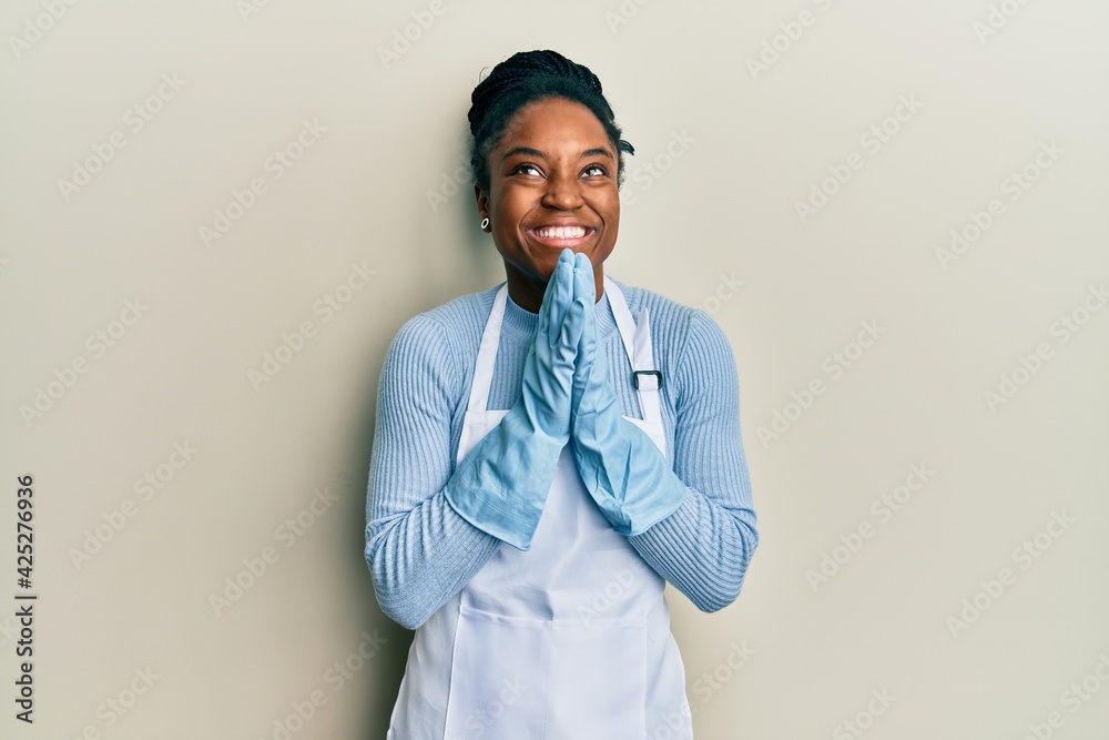 African american woman with braided hair wearing cleaner apron and gloves begging and praying with hands together with hope expression on face very emotional and worried. begging. African american woman with braided hair wearing cleaner apron and gloves begging and praying with hands together with hope expression on face very emotional and worried. begging.