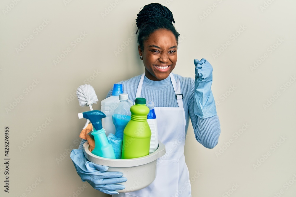 African american woman with braided hair wearing apron holding cleaning products screaming proud, celebrating victory and success very excited with raised arms African american woman with braided hair wearing apron holding cleaning products screaming proud, celebrating victory and success very excited with raised arms