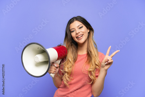 Young Russian girl isolated on blue background holding a megaphone and smiling and showing victory sign
