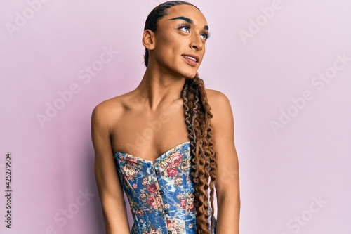 Hispanic man wearing make up and long hair wearing elegant corset looking away to side with smile on face, natural expression. laughing confident.