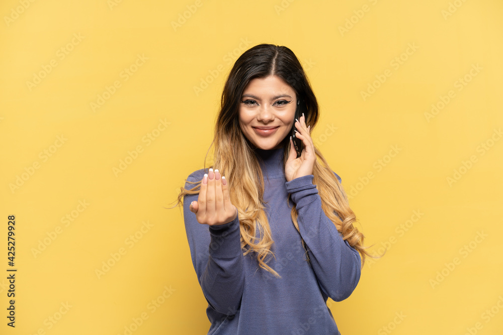 Young Russian girl using mobile phone isolated on yellow background inviting to come with hand. Happy that you came Young Russian girl using mobile phone isolated on yellow background inviting to come with hand. Happy that you came
