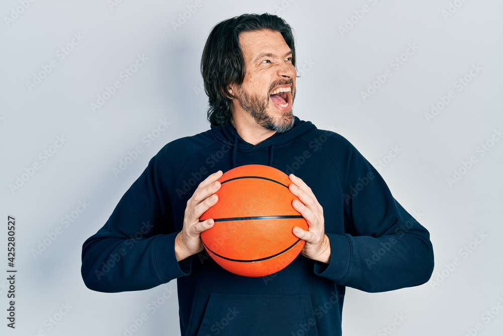 Middle age caucasian man holding basketball ball angry and mad screaming frustrated and furious, shouting with anger. rage and aggressive concept.