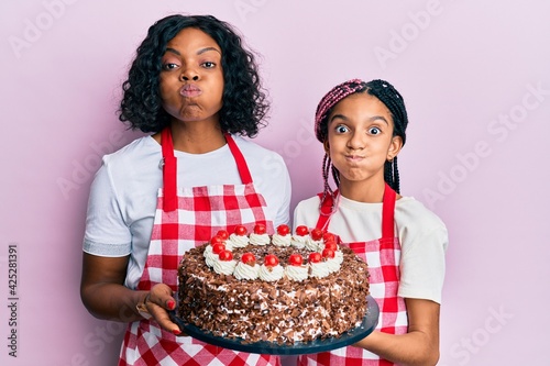 Beautiful african american mother and daughter wearing baker apron holding homemade cake puffing cheeks with funny face. mouth inflated with air, catching air.