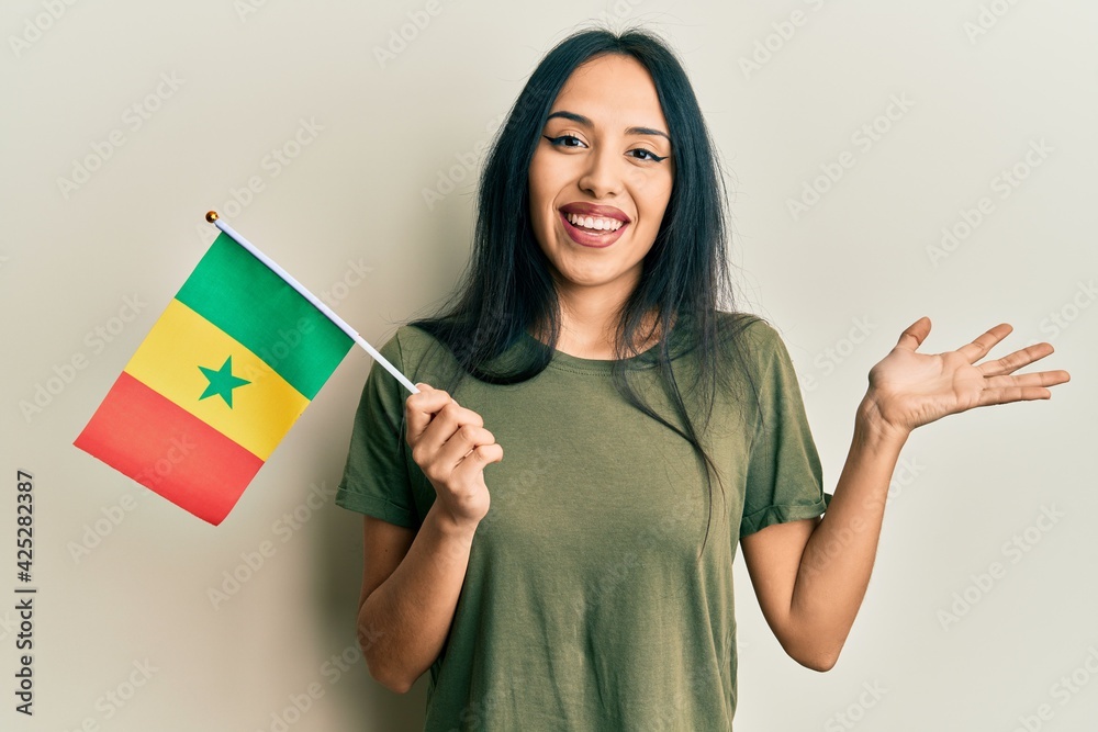 Young hispanic girl holding senegal flag celebrating achievement with happy smile and winner expression with raised hand Young hispanic girl holding senegal flag celebrating achievement with happy smile and winner expression with raised hand