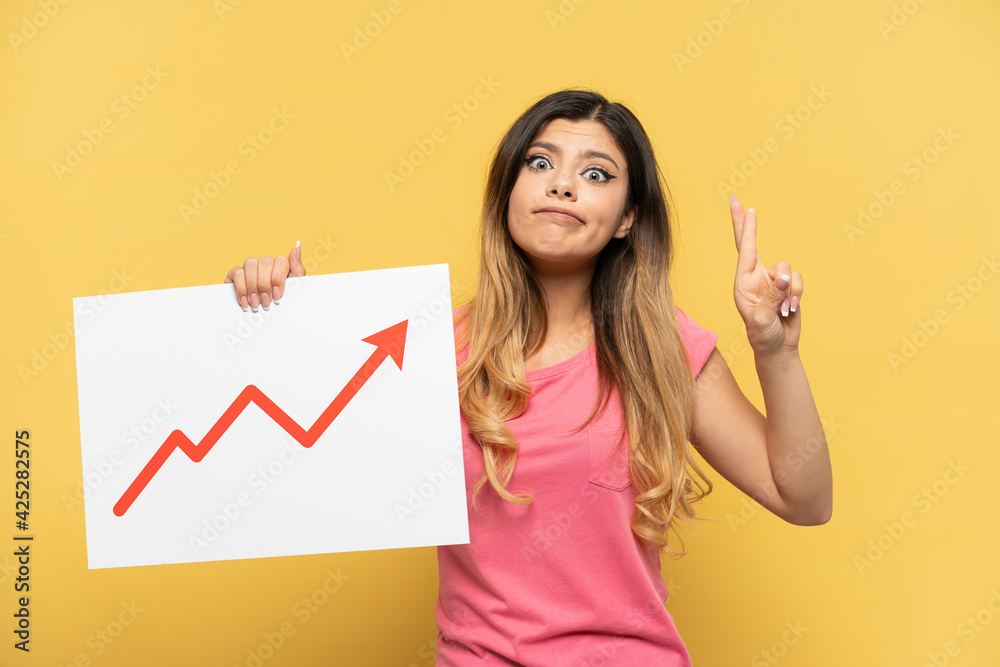 Young Russian girl isolated on yellow background holding a sign with a growing statistics arrow symbol with fingers crossing Young Russian girl isolated on yellow background holding a sign with a growing statistics arrow symbol with fingers crossing
