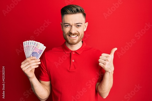 Young redhead man holding 20 polish zloty banknotes smiling happy and positive, thumb up doing excellent and approval sign