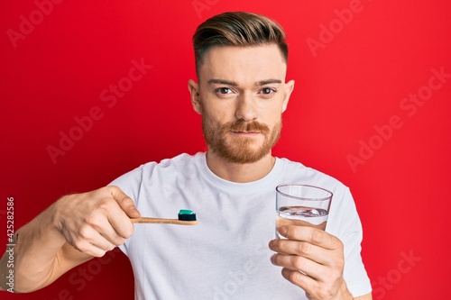 Young redhead man holding toothbrush with toothpaste and glass of water relaxed with serious expression on face. simple and natural looking at the camera.