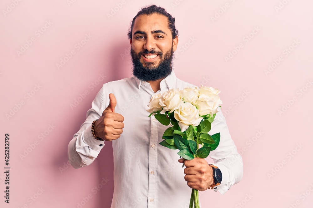 Young arab man holding flowers smiling happy and positive, thumb up doing excellent and approval sign Young arab man holding flowers smiling happy and positive, thumb up doing excellent and approval sign