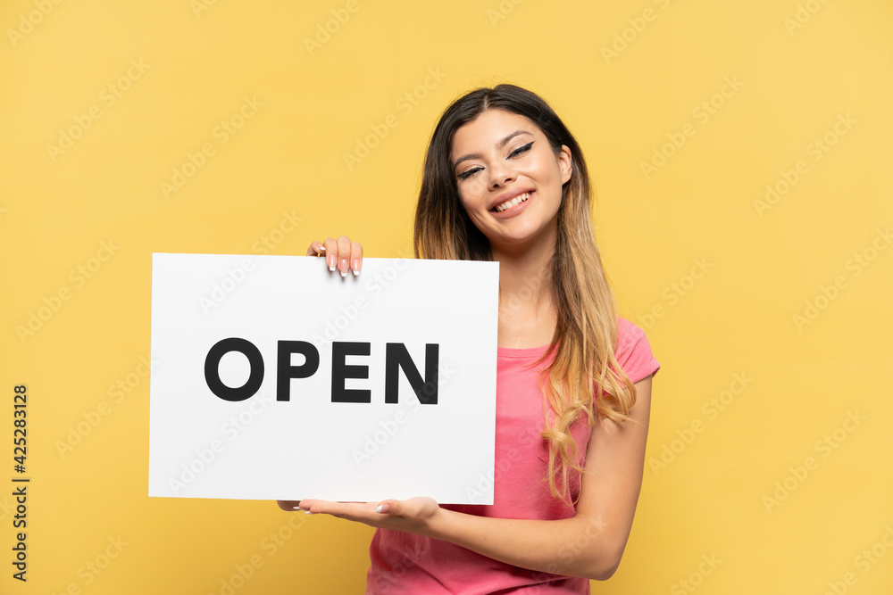 Young Russian girl isolated on yellow background holding a placard with text OPEN with happy expression Young Russian girl isolated on yellow background holding a placard with text OPEN with happy expression