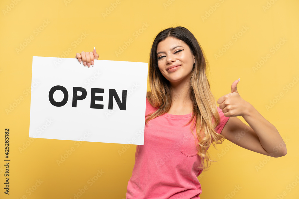 Young Russian girl isolated on yellow background holding a placard with text OPEN with thumb up Young Russian girl isolated on yellow background holding a placard with text OPEN with thumb up