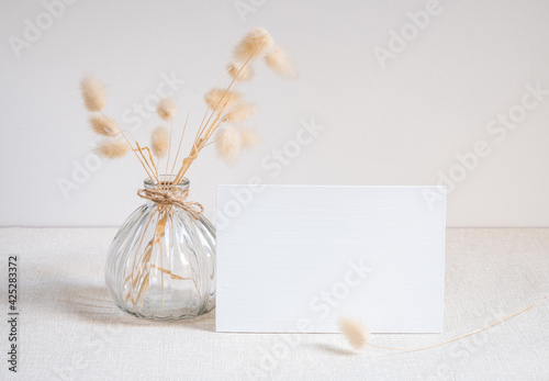 Blank white greeting card mock up.decoration with dried Lagurus ovatus flowers composition in modern glass vase  on  beige table and cement wall background