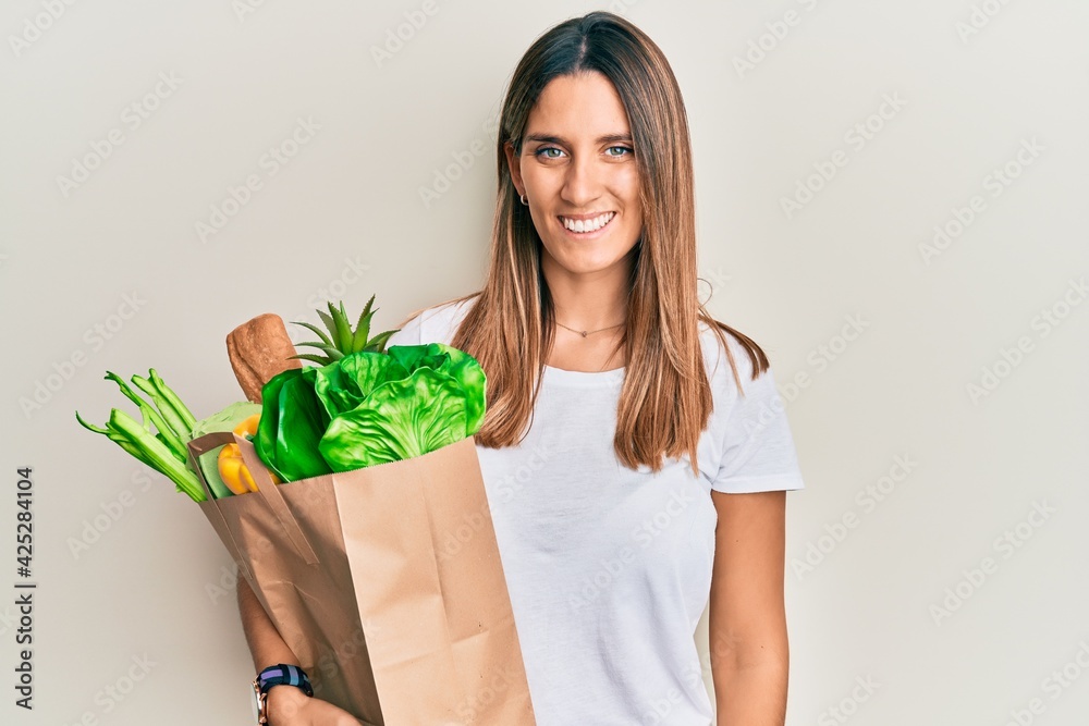 Brunette young woman holding paper bag with bread and groceries looking positive and happy standing and smiling with a confident smile showing teeth Brunette young woman holding paper bag with bread and groceries looking positive and happy standing and smiling with a confident smile showing teeth