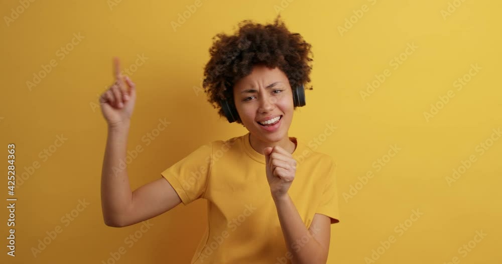 Positive young African American girl sings favorite song keeps hand near mouth pretends singing in microphone dressed casually isolated over yellow studio background. People lifestyle fun concept Positive young African American girl sings favorite song keeps hand near mouth pretends singing in microphone dressed casually isolated over yellow studio background. People lifestyle fun concept