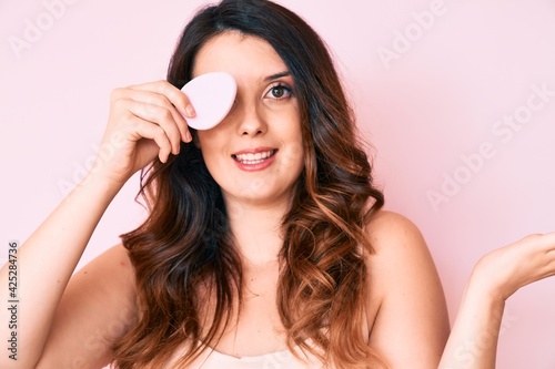 Young beautiful brunette woman holding makeup sponge celebrating achievement with happy smile and winner expression with raised hand