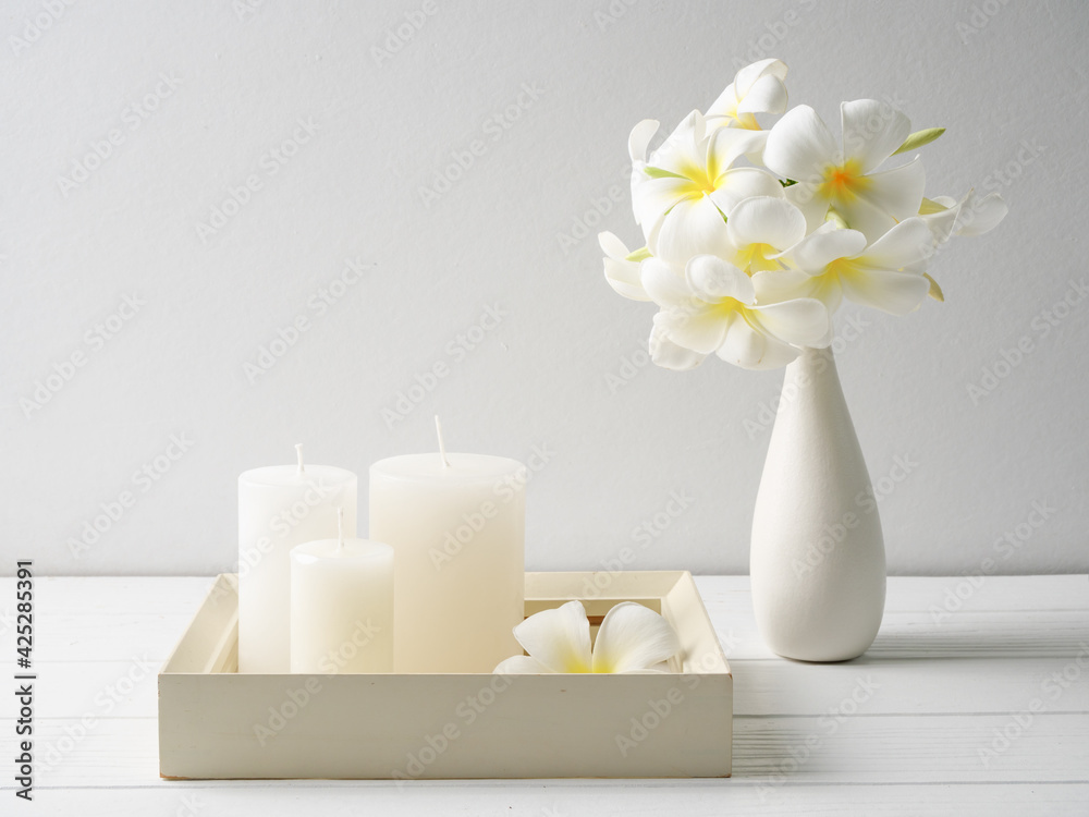 White candles on white wood tray and beautiful Plumeria flowers in modern glass vase composition over white wooden table room interior White candles on white wood tray and beautiful Plumeria flowers in modern glass vase composition over white wooden table room interior