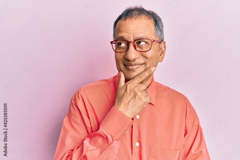 Middle age indian man wearing casual clothes and glasses with hand on chin thinking about question, pensive expression. smiling with thoughtful face. doubt concept. Middle age indian man wearing casual clothes and glasses with hand on chin thinking about question, pensive expression. smiling with thoughtful face. doubt concept.