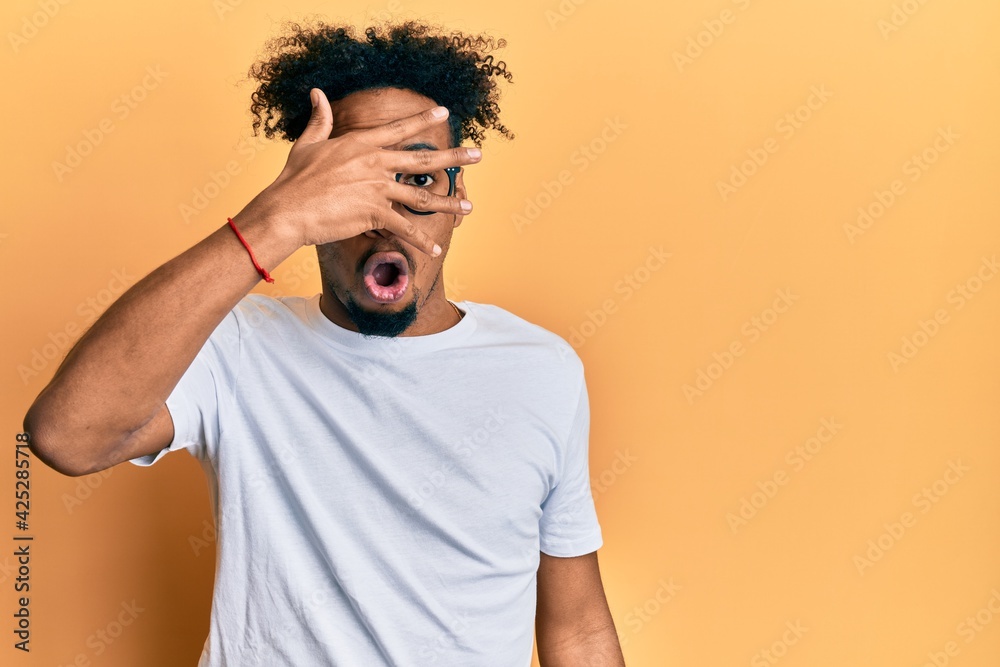 Young african american man with beard wearing casual white t shirt and glasses peeking in shock covering face and eyes with hand, looking through fingers afraid Young african american man with beard wearing casual white t shirt and glasses peeking in shock covering face and eyes with hand, looking through fingers afraid