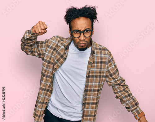 Young african american man with beard wearing casual clothes and glasses angry and mad raising fist frustrated and furious while shouting with anger. rage and aggressive concept.
