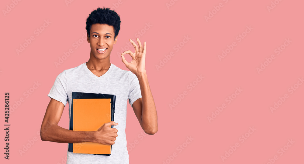 Young african american man holding book doing ok sign with fingers, smiling friendly gesturing excellent symbol