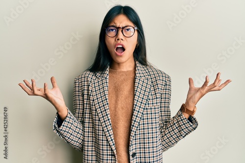Young chinese woman wearing business style and glasses crazy and mad shouting and yelling with aggressive expression and arms raised. frustration concept.