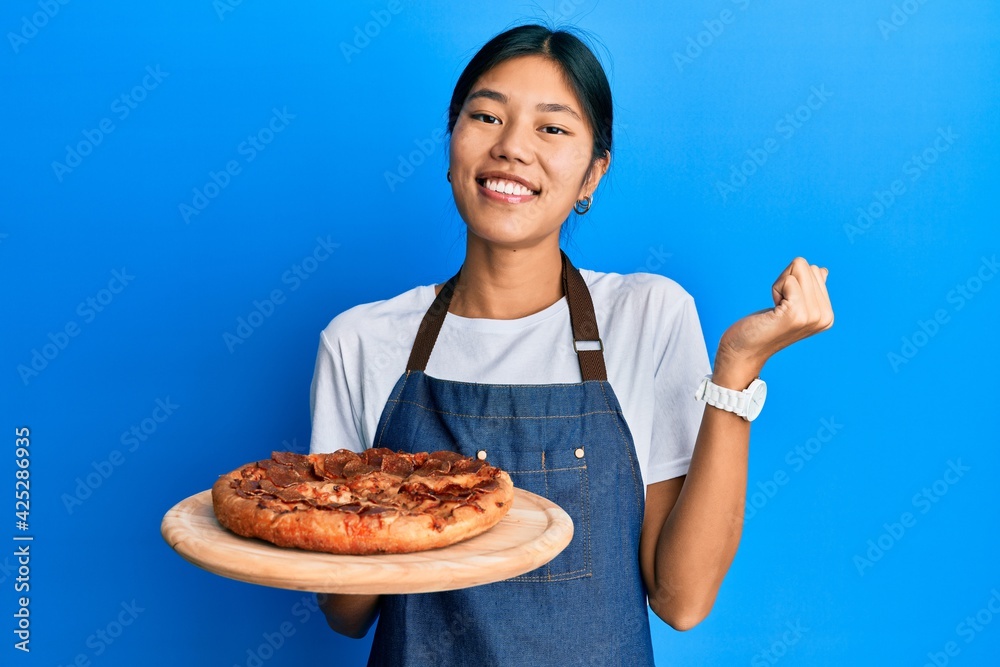 Young chinese woman wearing waiter apron holding italian pizza screaming proud, celebrating victory and success very excited with raised arm Young chinese woman wearing waiter apron holding italian pizza screaming proud, celebrating victory and success very excited with raised arm