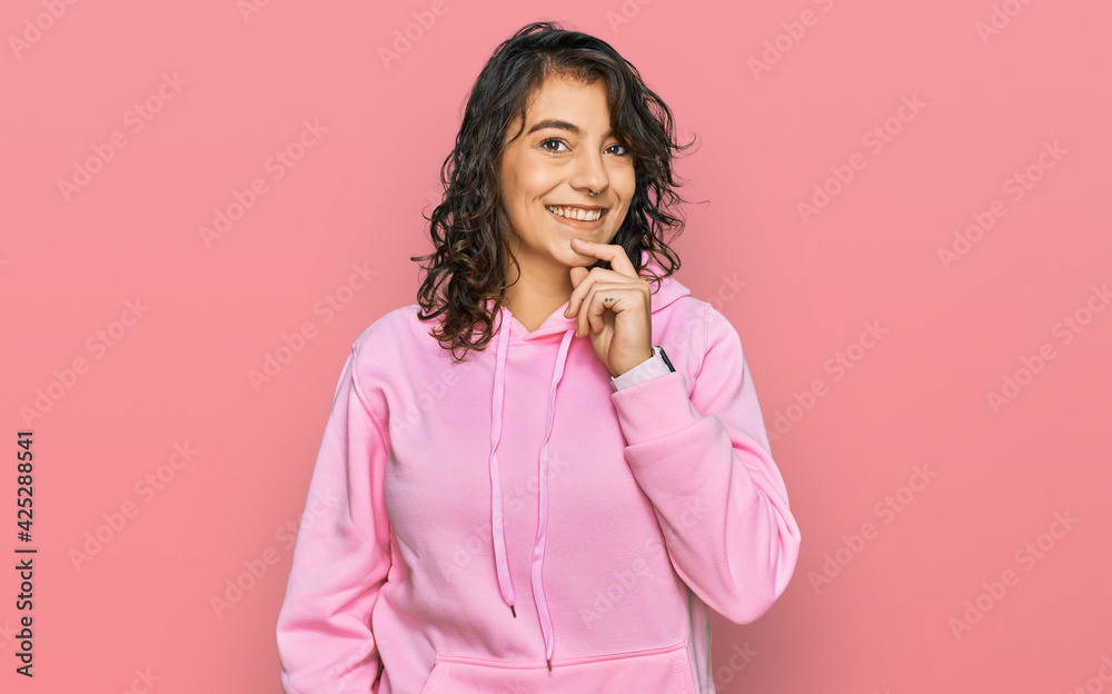 Young hispanic woman wearing casual sweatshirt looking confident at the camera smiling with crossed arms and hand raised on chin. thinking positive. Young hispanic woman wearing casual sweatshirt looking confident at the camera smiling with crossed arms and hand raised on chin. thinking positive.