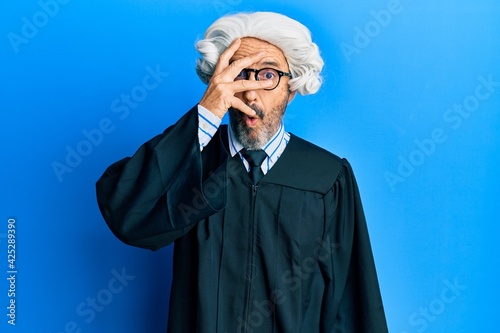 Middle age hispanic man wearing judge uniform peeking in shock covering face and eyes with hand, looking through fingers afraid