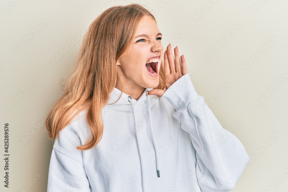 Beautiful young caucasian girl wearing casual sweatshirt shouting and screaming loud to side with hand on mouth. communication concept. Beautiful young caucasian girl wearing casual sweatshirt shouting and screaming loud to side with hand on mouth. communication concept.