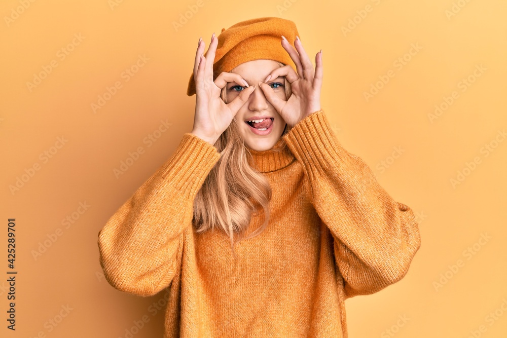 Beautiful young caucasian girl wearing french look with beret doing ok gesture like binoculars sticking tongue out, eyes looking through fingers. crazy expression. Beautiful young caucasian girl wearing french look with beret doing ok gesture like binoculars sticking tongue out, eyes looking through fingers. crazy expression.