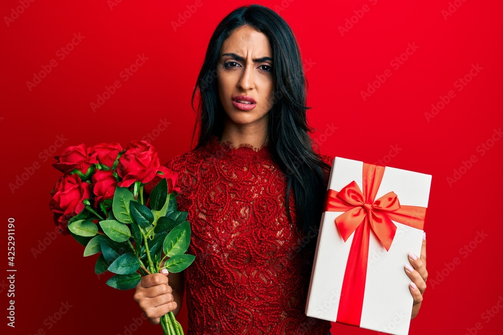 Beautiful hispanic woman holding anniversary gift and roses bouquet clueless and confused expression. doubt concept. Beautiful hispanic woman holding anniversary gift and roses bouquet clueless and confused expression. doubt concept.