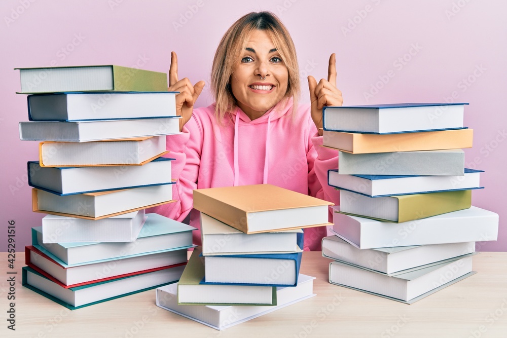 Young caucasian woman sitting on the table with books smiling amazed and surprised and pointing up with fingers and raised arms. Young caucasian woman sitting on the table with books smiling amazed and surprised and pointing up with fingers and raised arms.