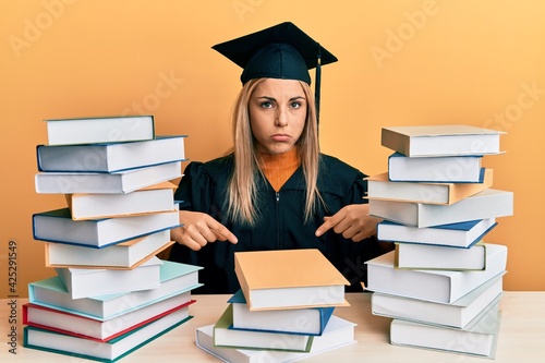 Young caucasian woman wearing graduation ceremony robe sitting on the table pointing down looking sad and upset, indicating direction with fingers, unhappy and depressed.