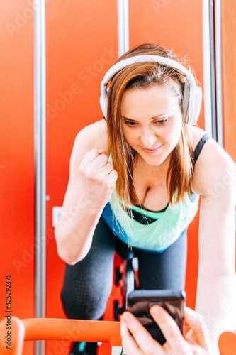 Foto Young fit athlete woman looking at smart phone while doing biking indor clenching her fist