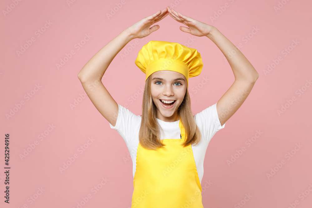 Teen happy girl chef cook confectioner baker housewife in yellow apron white t-shirt cap holding folded hand above head like house roof stay home isolated on pink background studio. Food cake concept. Teen happy girl chef cook confectioner baker housewife in yellow apron white t-shirt cap holding folded hand above head like house roof stay home isolated on pink background studio. Food cake concept.