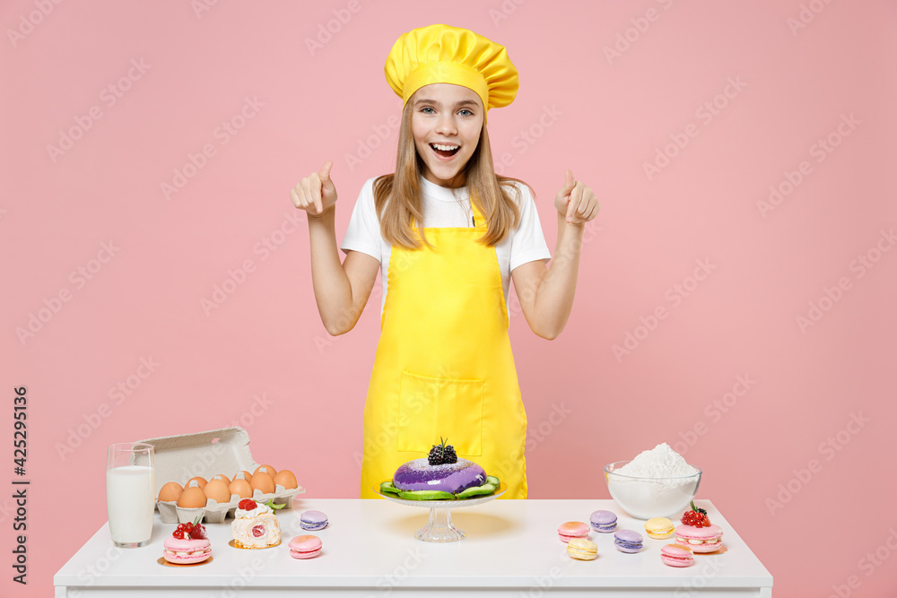 Teen smiling girl chef cook confectioner baker in yellow apron cap point index finger down on table workspace mock up area isolated on pastel pink background Mousse cake food process workshop concept Teen smiling girl chef cook confectioner baker in yellow apron cap point index finger down on table workspace mock up area isolated on pastel pink background Mousse cake food process workshop concept